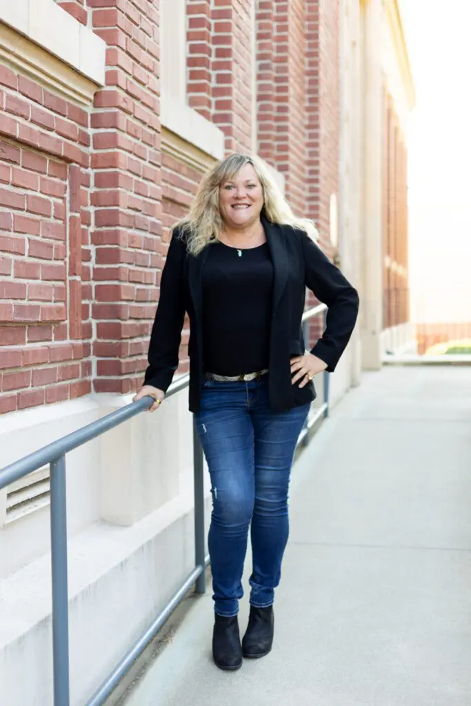 A woman with blond hair, a writer and consultant for entrepreneurs, stands outdoors by a brick building in a black blazer, jeans, and boots, holding a metal railing and smiling at the camera.
