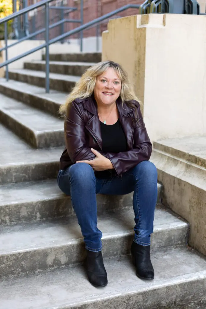 Seattle writer and consultant with blonde hair wearing a maroon leather jacket, black top, and jeans, sits smiling on outdoor concrete steps.