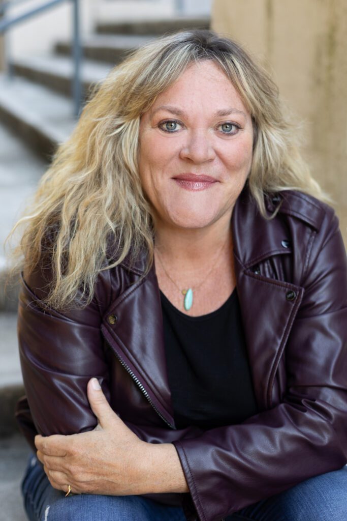 A woman with wavy blonde hair wearing a maroon leather jacket and black shirt sits on outdoor steps, looking at the camera with a slight smile.