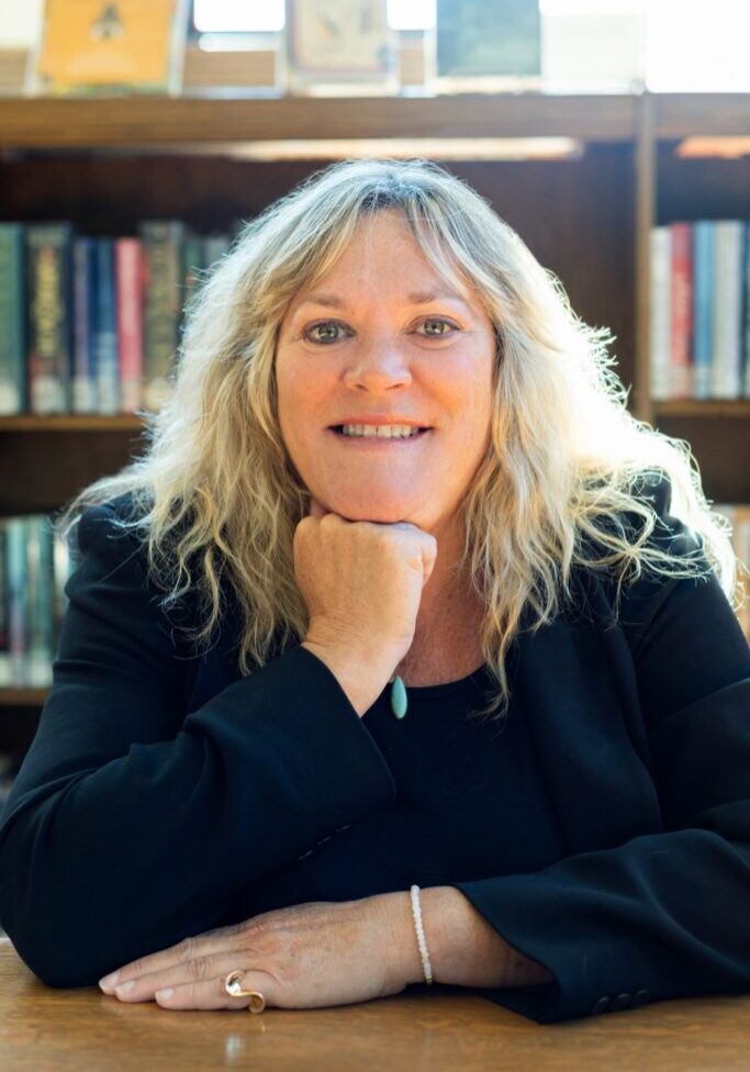 A woman with long blonde hair sits at a wooden table, resting her chin on her hand, and smiling in front of bookshelves in a well-lit library—capturing the confidence of a skilled business launch consultant.